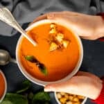 Woman hands holding a bowl of tomato soup topped with croutons and fresh basil and a spoon inside it.