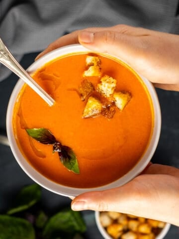 Woman hands holding a bowl of tomato soup topped with croutons and fresh basil and a spoon inside it.