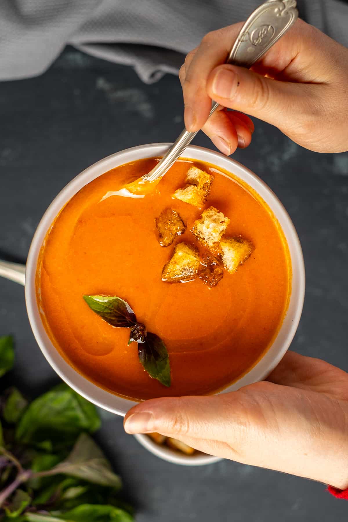 A hand holding a bowl of tomato soup and another hand holding the spoon inside it.