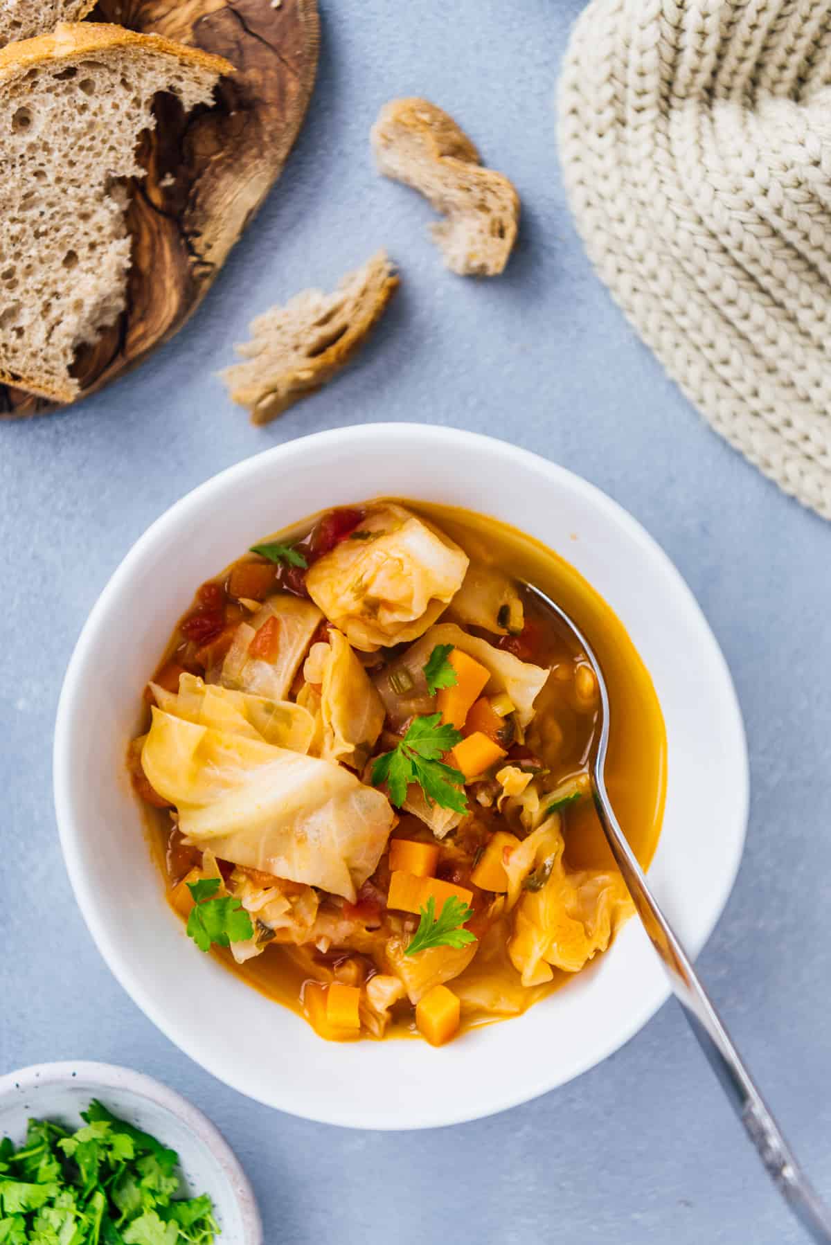 Vegetarian cabbage soup with carrots and parsley in a ceramic bowl and a spoon inside it. Bread slices, parsley and a knitted scarf on the side.
