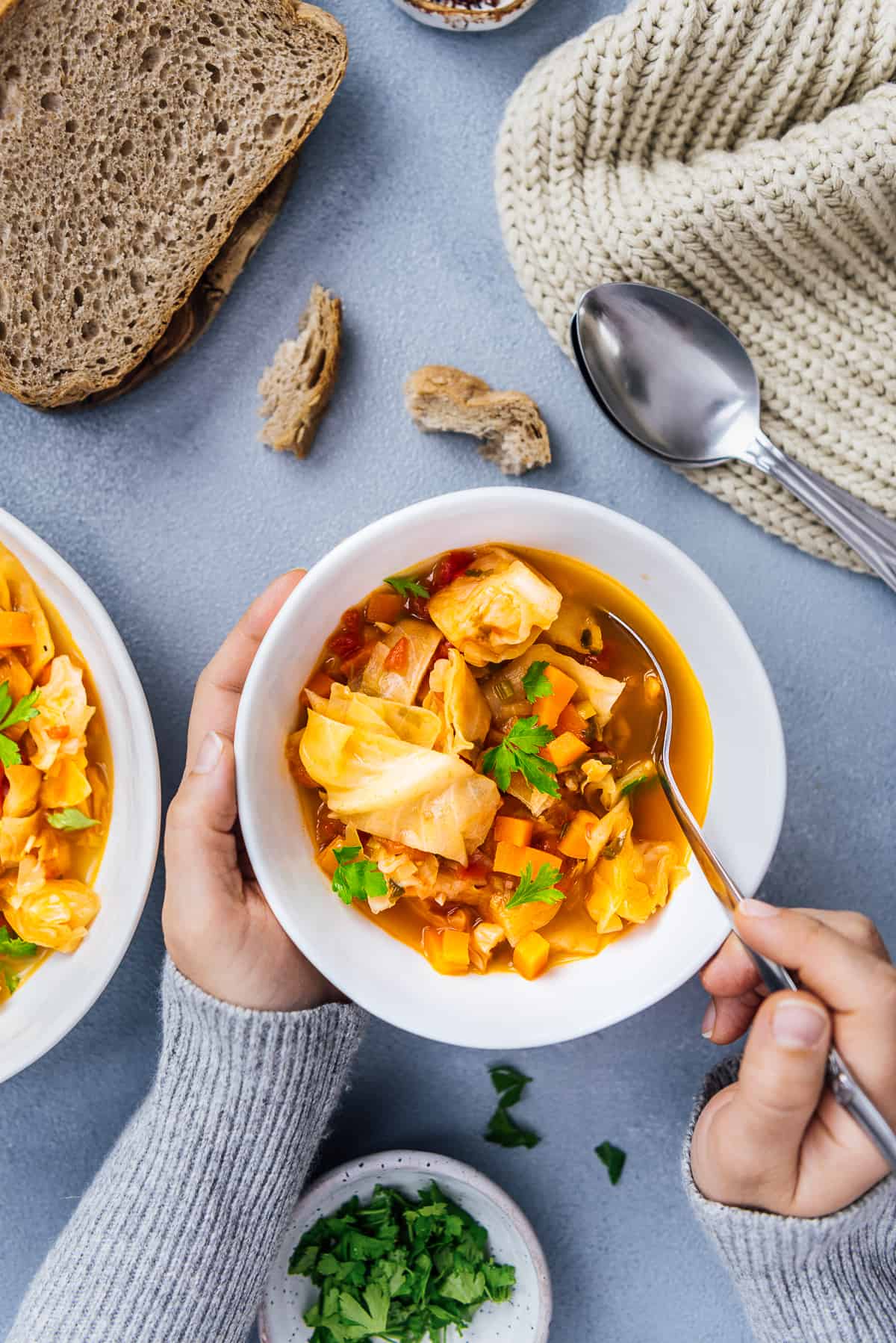 Woman having cabbage soup in a white bowl, another bowl of soup, bread slices, chili flakes in a small ceramic bowl, parsley and spoons on the side.