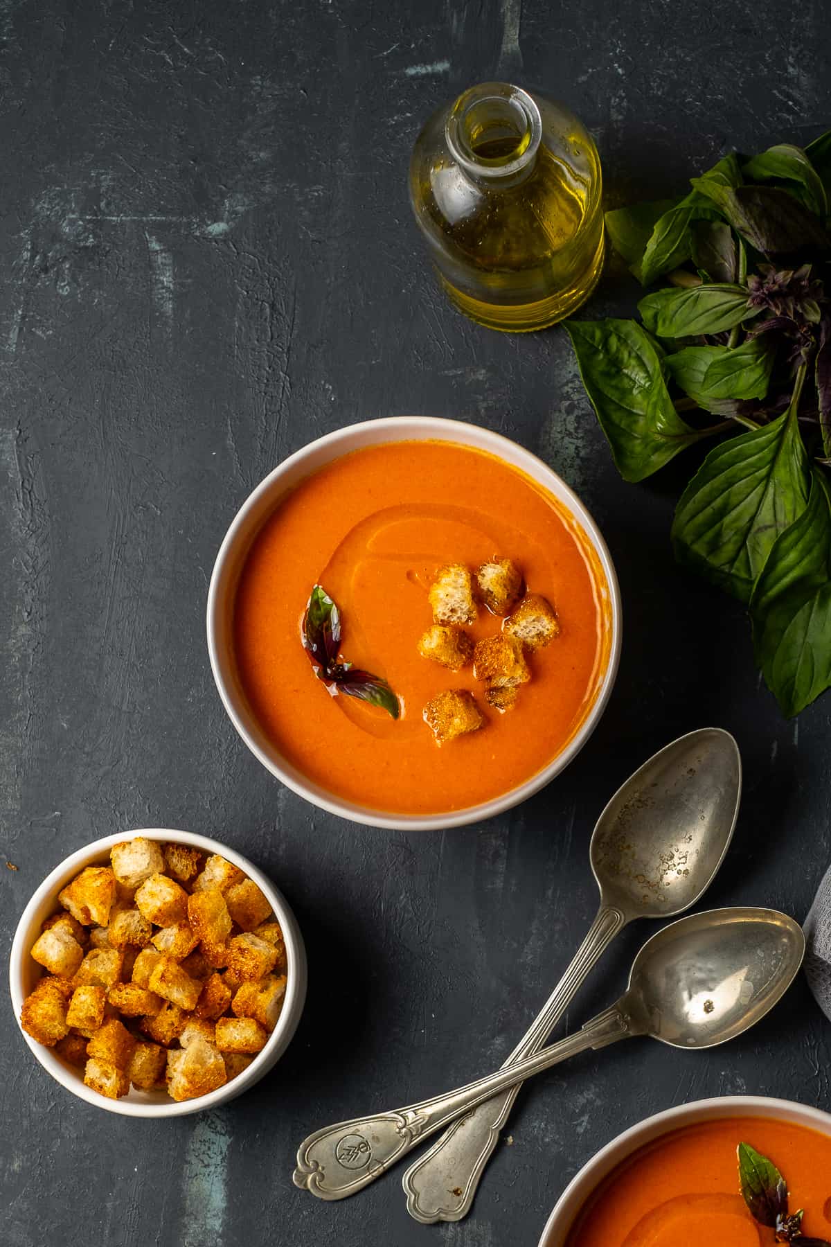 A bowl of tomato soup on a dark backdrop, vintage spoons, croutons in a bowl and fresh basil on the side.