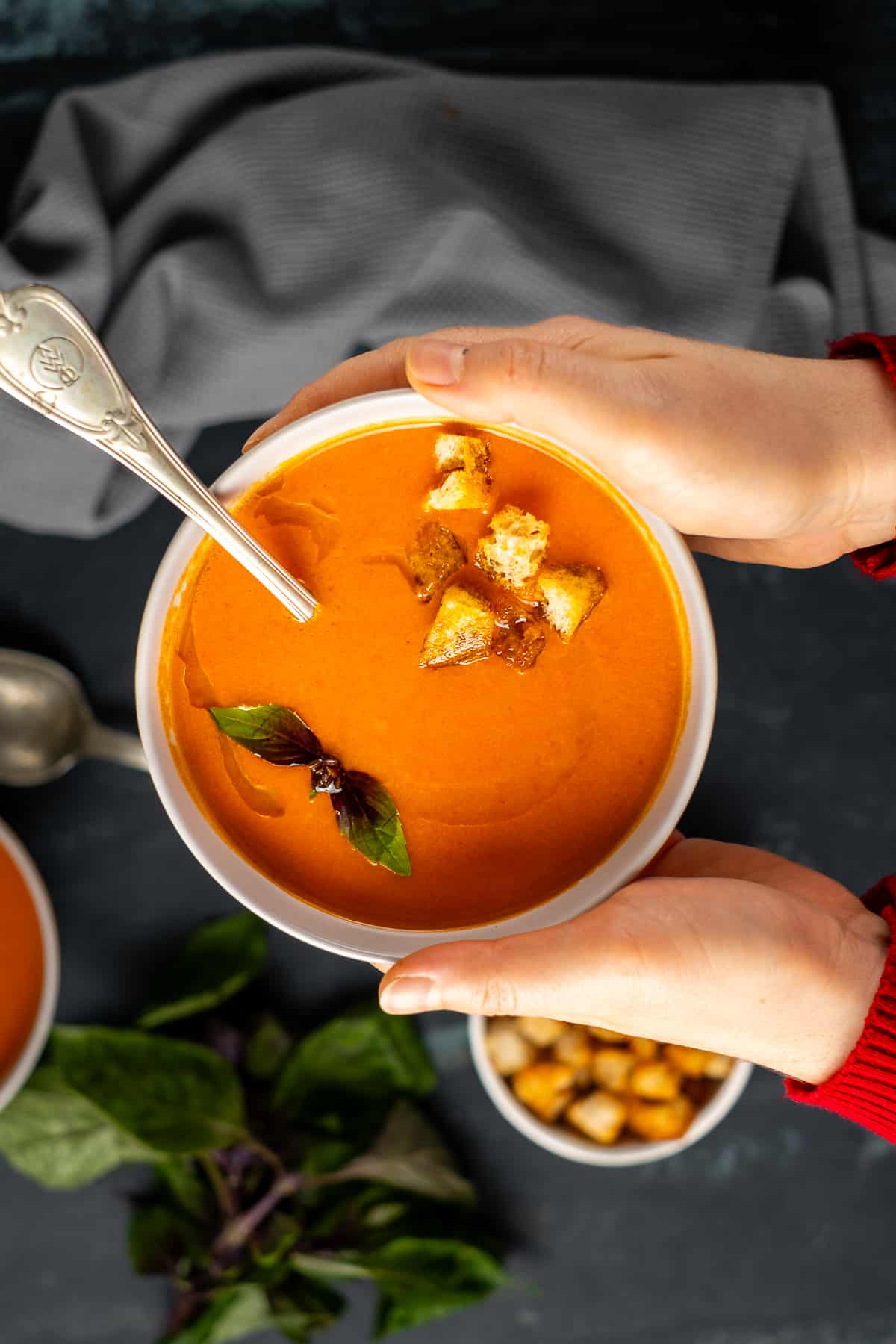Hands holding a bowl of tomato soup garnished with croutons and fresh basil.