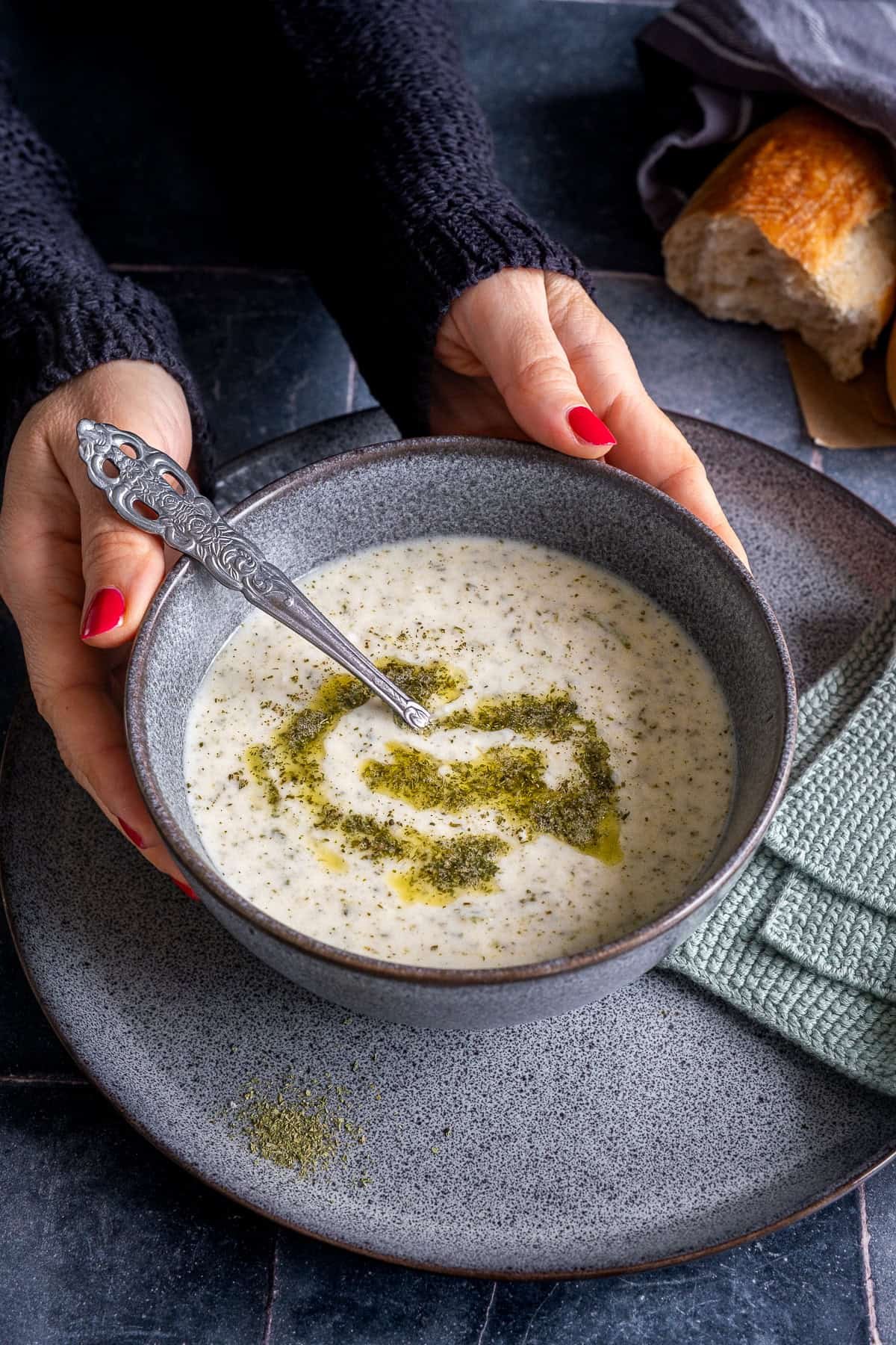 Hands holding a bowl of Turkish Yogurt Soup (Yayla Çorbası) topped with butter-mint sauce, served with bread in the background.