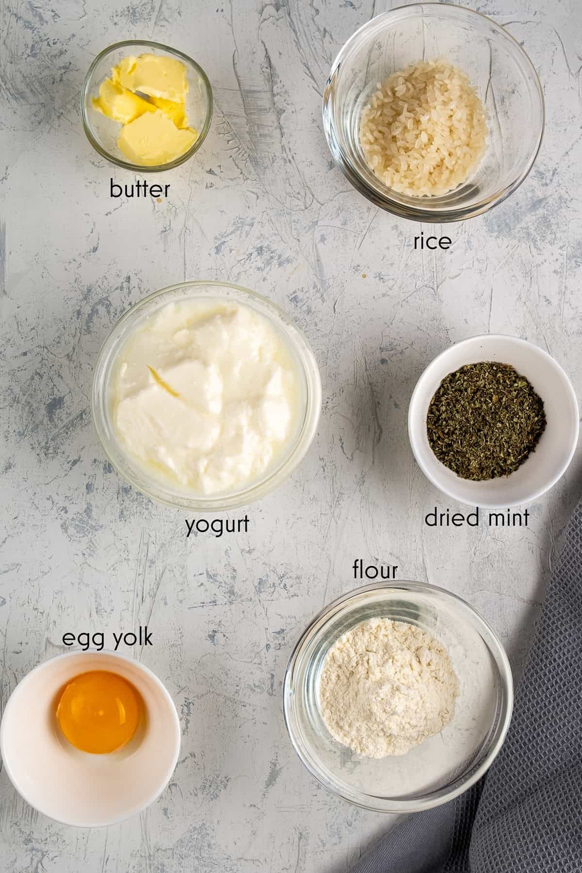 Ingredients for Turkish Yogurt Soup (Yayla Çorbası) arranged in bowls: yogurt, rice, flour, egg yolk, butter, and dried mint.