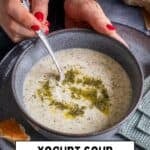 Hands holding a bowl of Turkish Yogurt Soup (Yayla Çorbası) topped with butter-mint sauce, served with bread in the background.