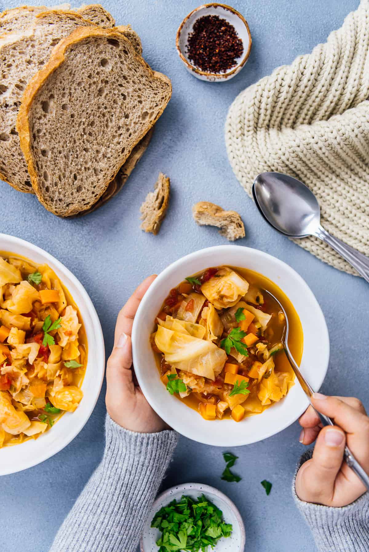 Woman hands with a light grey jumper holding a bowl of cabbage soup and a spoon. Bread slices, another bowl of soup and a knitted scarf on the side.