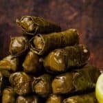 Stacked stuffed grape leaves (yaprak sarma) on a plate, showing tightly rolled, glossy leaves with a lemon wedge on the side against a dark background.