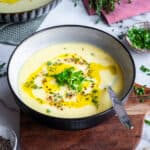 Celeriac soup in a bowl, topped with a drizzle of olive oil, black pepper, chopped parsley and thyme, and a spoon inside it.