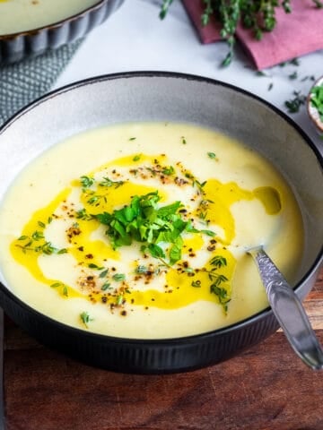 Celeriac soup in a bowl, topped with a drizzle of olive oil, black pepper, chopped parsley and thyme, and a spoon inside it.