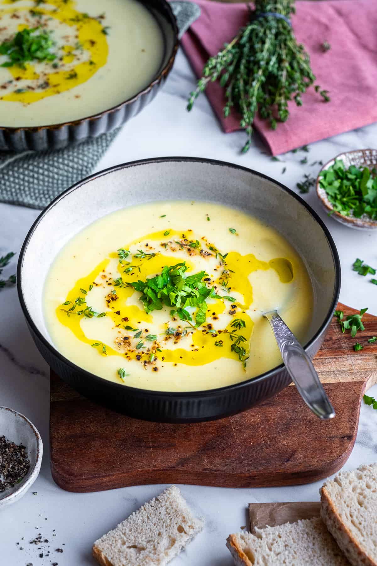 A bowl of celeriac soup garnished with a drizzle of olive oil, black pepper, chopped parsley and thyme, and a spoon in the bowl.