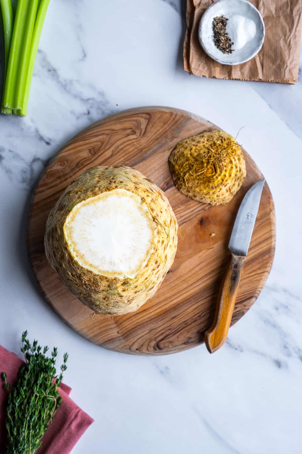 A celeriac vegetable on a wooden cutting board, its top cut off and a knife on the side.