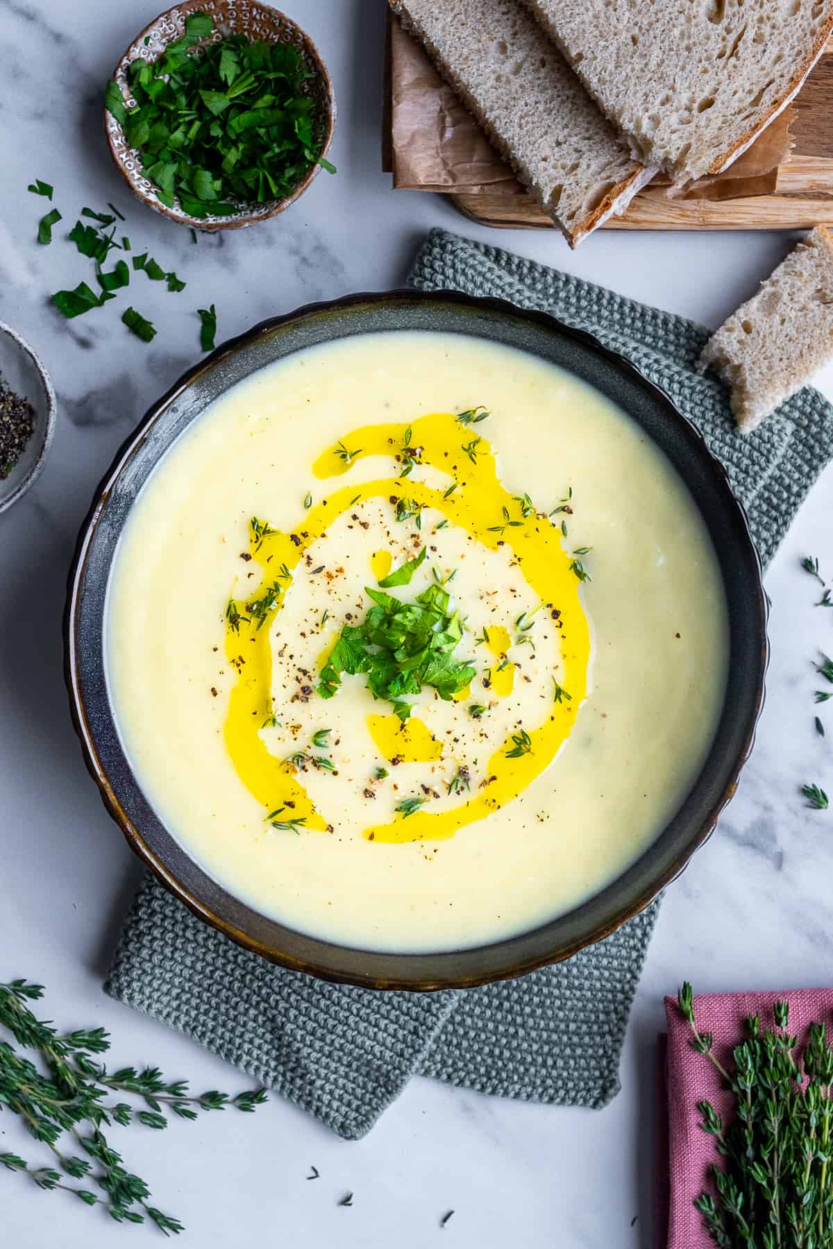 Creamy celeriac soup garnished with a drizzle of olive oil, black pepper, chopped parsley and thyme in a black bowl on a knitted napkin.