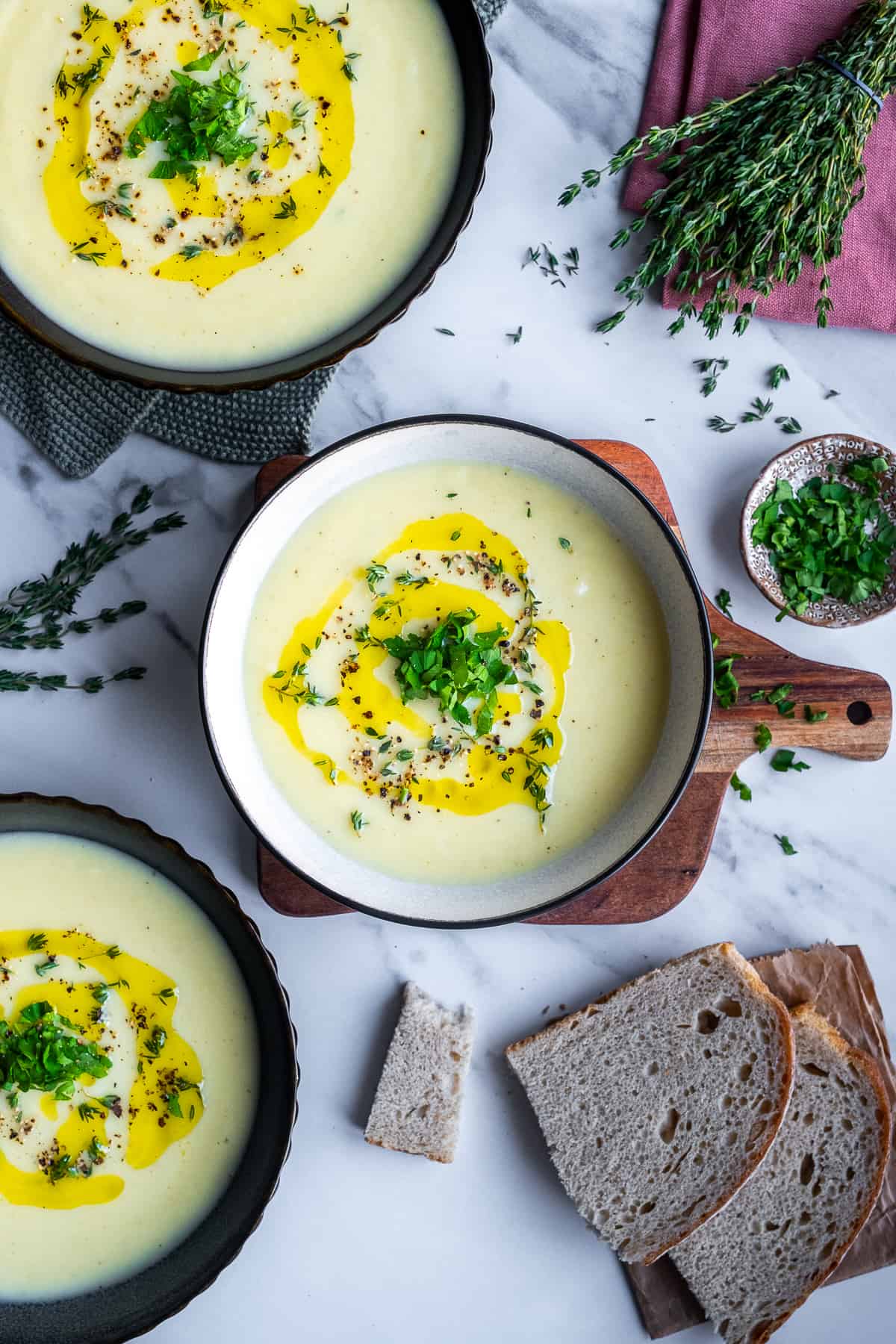 Three bowls of celeriac soup garnished with a drizzle of olive oil, chopped parsley and thyme, bread slices on the side.