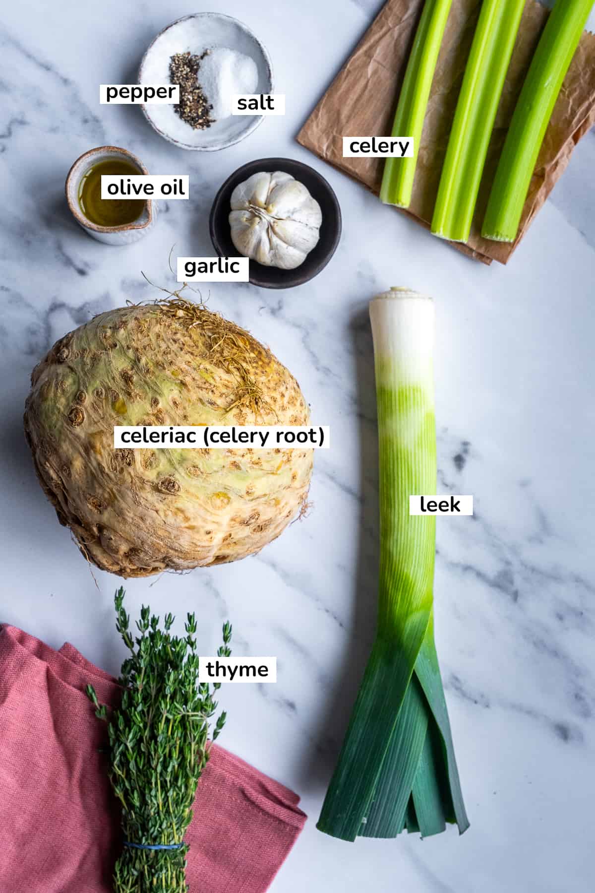 A celery root, leek, celery stalks, garlic, fresh thyme, olive oil, salt and pepper on a white marble.