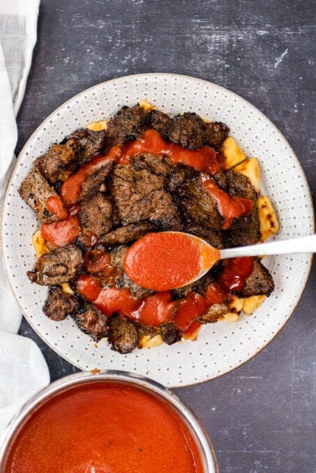 A spoon drizzling tomato sauce over cooked iskender meat over pide bread on a white plate.