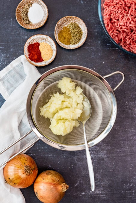 Pureed onion in a sieve for its juice, two large onions, spices in mini bowls and ground beef on the side on a dark background.