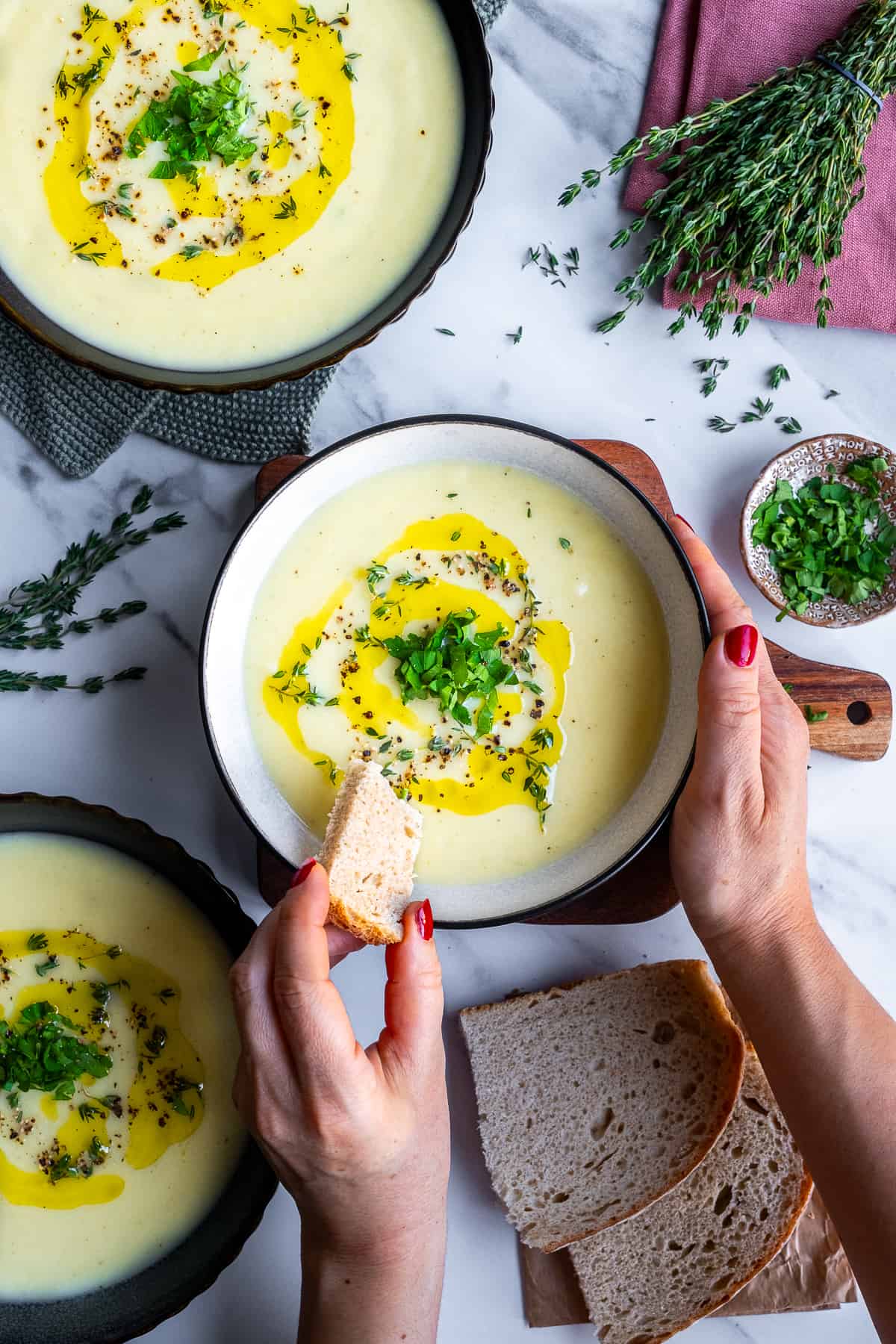 Woman about to dip a piece of bread into celeriac soup in a bowl, more soup bowls, fresh parsley and thyme on the side.