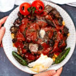 Hands holding homemade iskender kebab with tomato sauce, yogurt, roasted tomatoes and pepper on a white plate.