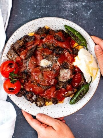 Hands holding a white plate with homemade iskender kebab with pide bread, tomato sauce, browned butter, yogurt, roasted tomatoes and green peppers.