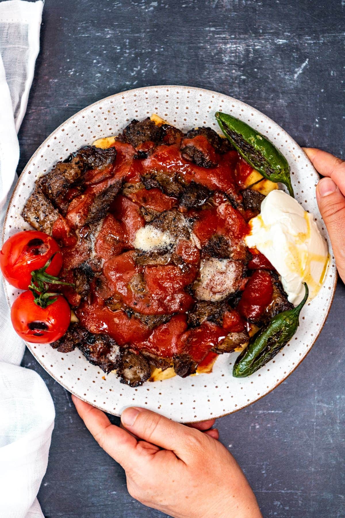 Hands holding a white plate with homemade iskender kebab with pide bread, tomato sauce, browned butter, yogurt, roasted tomatoes and green peppers.