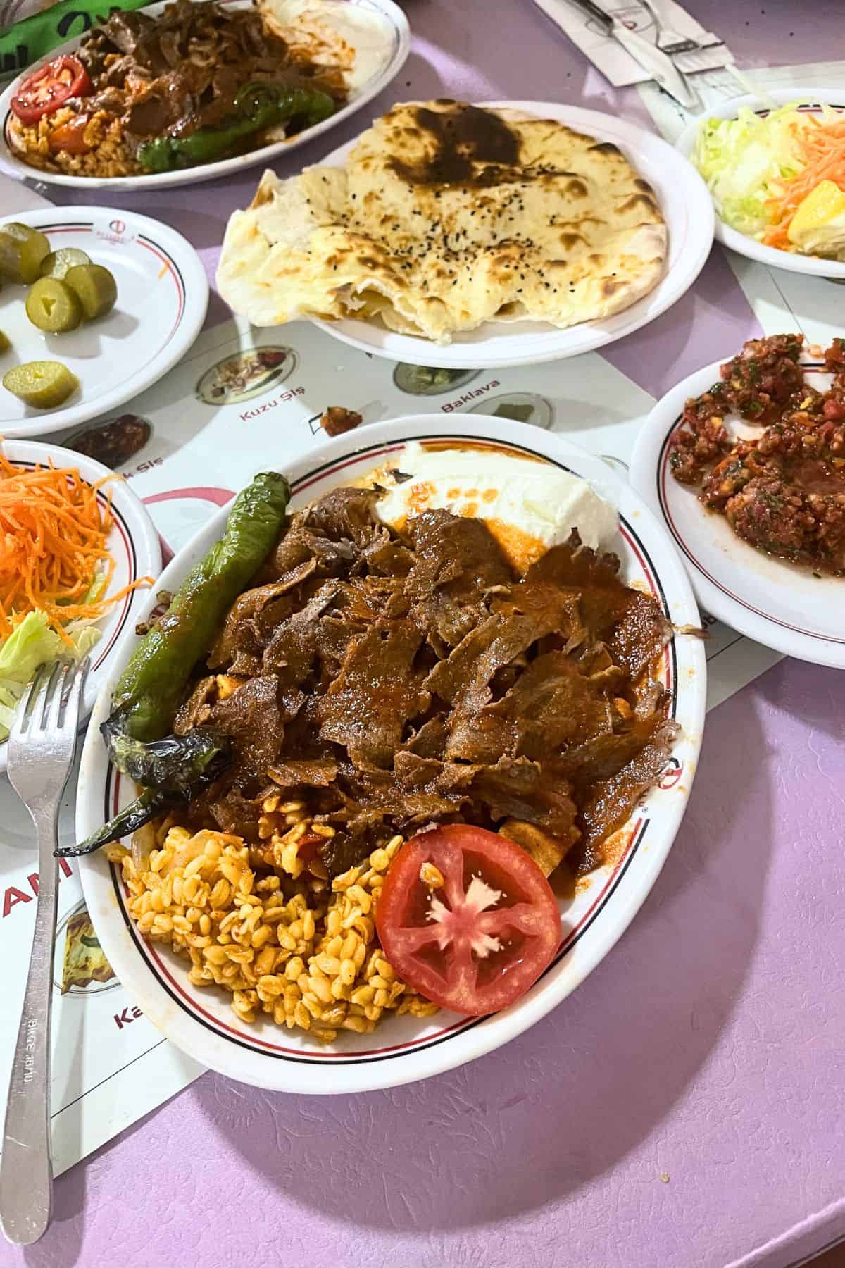 Restaurant-style iskender kebab served with sliced döner meat over bread, yogurt, tomato sauce, grilled green pepper, bulgur pilaf, and fresh tomato, alongside Turkish sides and flatbread.