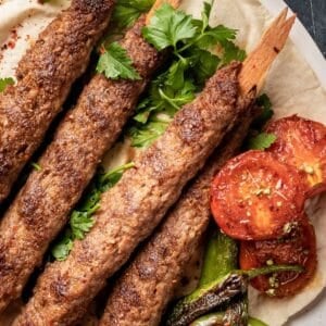 Adana kebabs on wooden skewers served on a flatbread, garnished with grilled tomatoes and peppers.