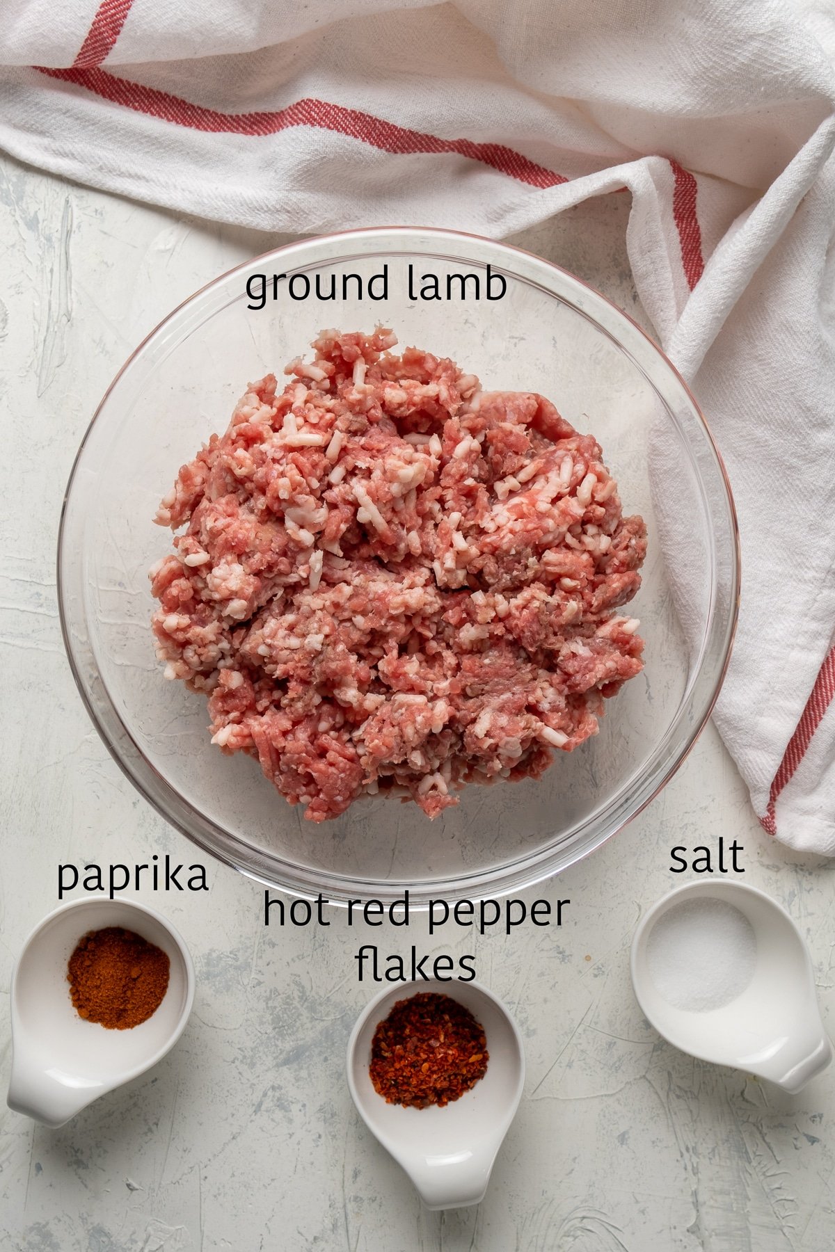 Ground lamb in a glass bowl, paprika, red pepper flakes and salt in separate bowls on the side.