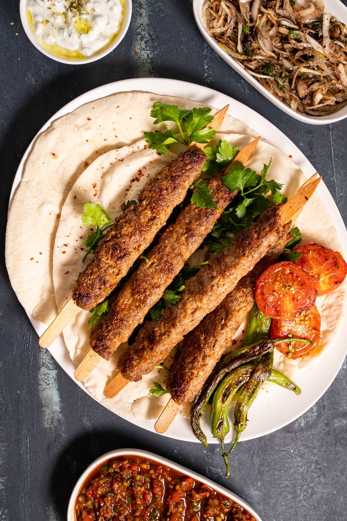 Ground lamb kebabs served on lavash with roasted tomatoes and green peppers on a white plate, ezme and sumac onions on the side.