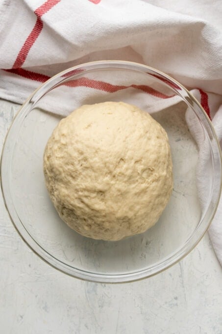 Bread dough in a glass mixing bowl, a white kitchen towel is on the side.