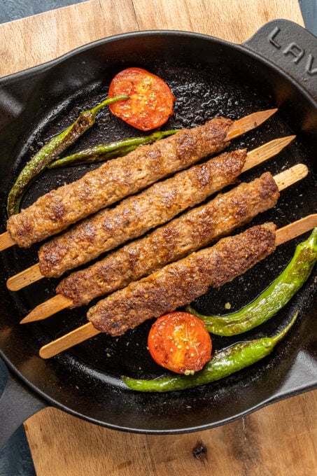 Minced lamb kebabs in an iron skillet and tomatoes and green peppers inside it.