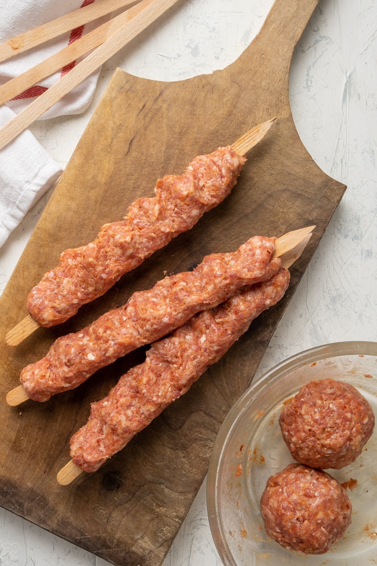 Spicy ground lamb mixture shaped on three wooden skewers on a wooden cutting board and two large balls of meat in a bowl on the side.