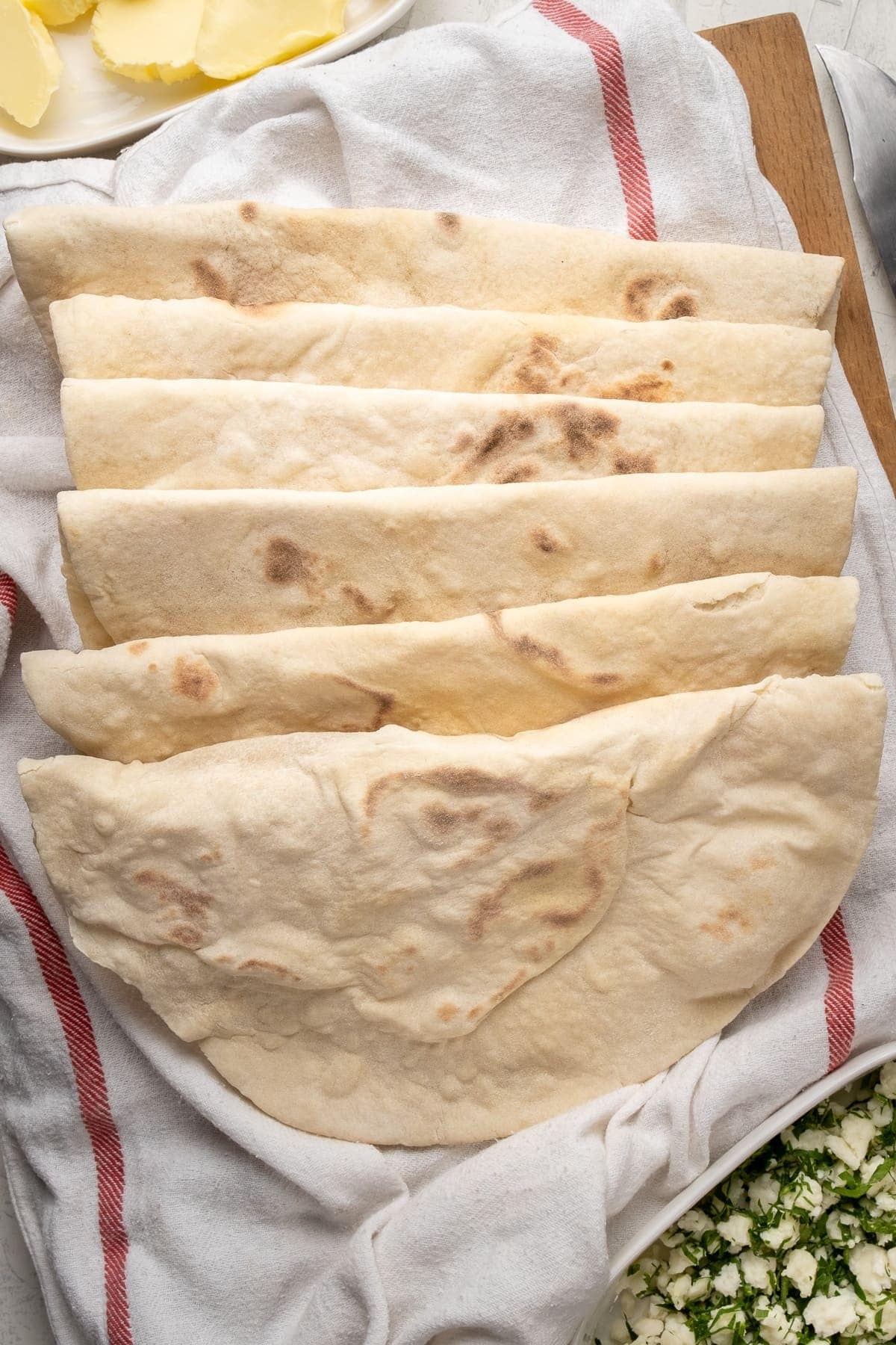 Lavash breads on a clean white kitchen towel. Butter on a white oval plate on the top and parsley and feta on another white oval plate at the bottom.