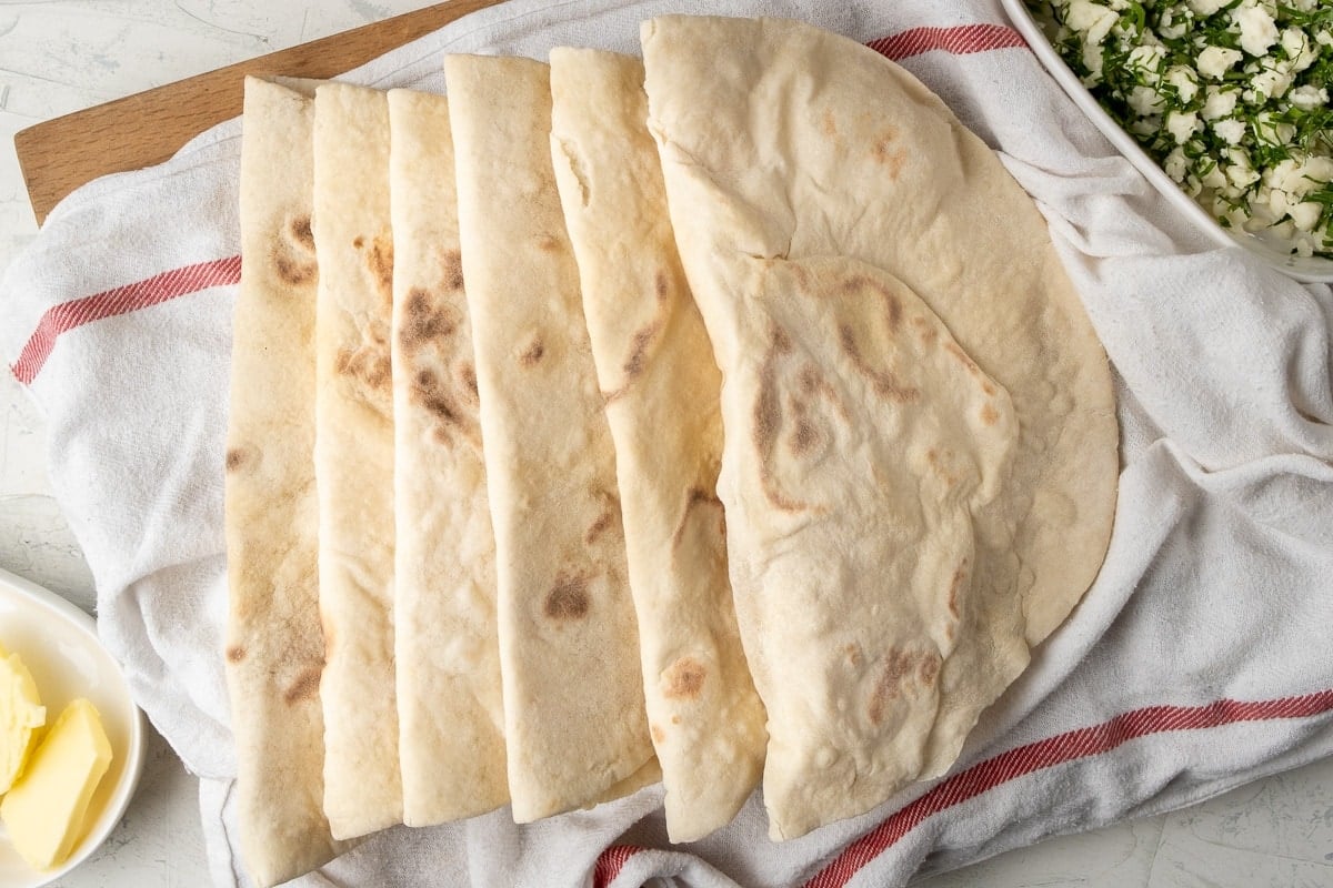 Lavash flat breads on a white kitchen towel and some butter in a small oval bowl on one side and feta parsley mixture in another oval bowl on the other side of the breads.