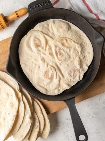 Lavash breads in an iron skillet on a wooden board and more lavash breads on the side.