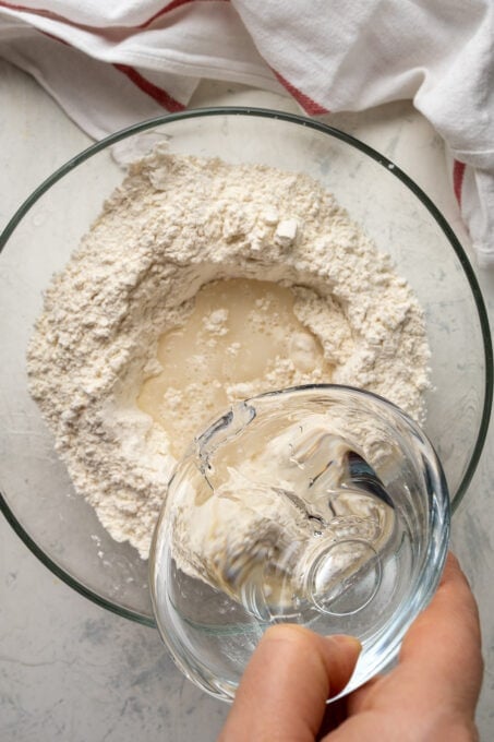 A hand pouring water into the flour mixture in a glass mixing bowl.