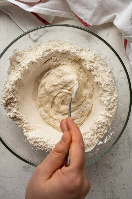 A hand mixing the flour and yeast mixture with water using a fork.