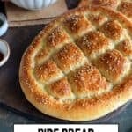 Freshly baked Turkish Pide Bread with a golden crust and sesame topping, served with a bowl of Turkish soup. Hands are seen holding the soup bowl in the background.