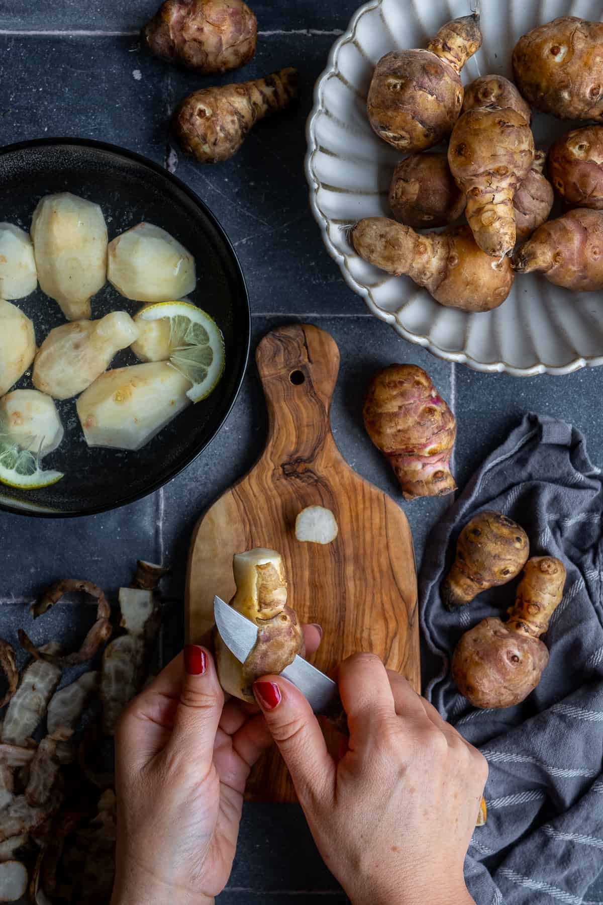 A Jerusalem artichoke being peeled with a knife, peeled and unpeeled Jerusalem artichokes on the side.