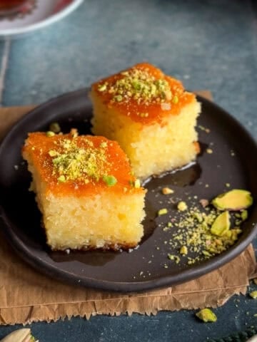 Two slices of Turkish revani semolina cake soaked in sweet syrup, topped with crushed pistachios and served on a dark plate.