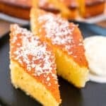Close-up of two slices of revani (Turkish semolina cake) on a black plate, golden and syrup-soaked, topped with desiccated coconut and served with a scoop of cream on the side.