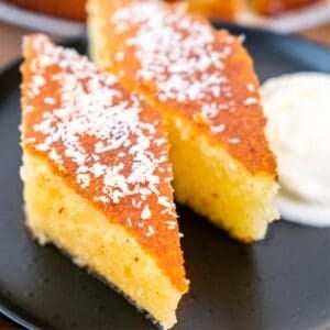 Close-up of two slices of revani (Turkish semolina cake) on a black plate, golden and syrup-soaked, topped with desiccated coconut and served with a scoop of cream on the side.