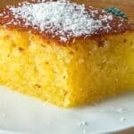 Close-up of a slice of revani (Turkish semolina cake) on a white plate, golden and syrup-soaked with a moist crumb, topped with desiccated coconut.