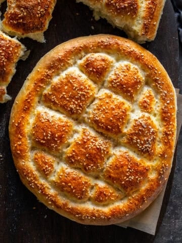 Golden Turkish Pide Bread with sesame seeds and a traditional diamond pattern, placed on a dark wooden surface. Another torn bread is visible in the background.