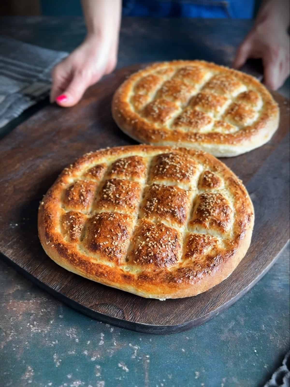 Freshly baked Turkish Pide Bread with a golden crust, sesame topping, and a traditional pattern, placed on a wooden board. Hands are seen in the background.