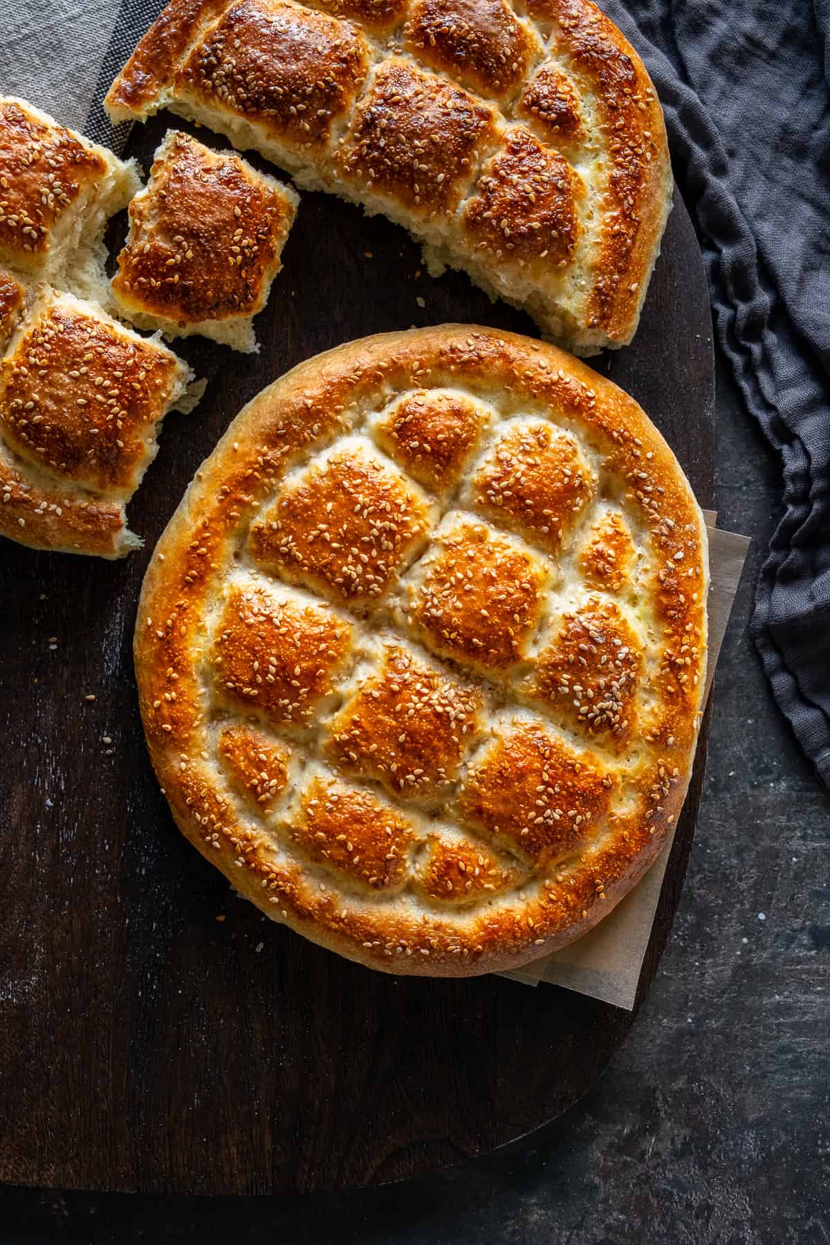 Freshly baked Turkish Pide Bread with a golden crust, sesame seeds, and a traditional diamond pattern, placed on a wooden surface. A torn loaf is visible in the background.