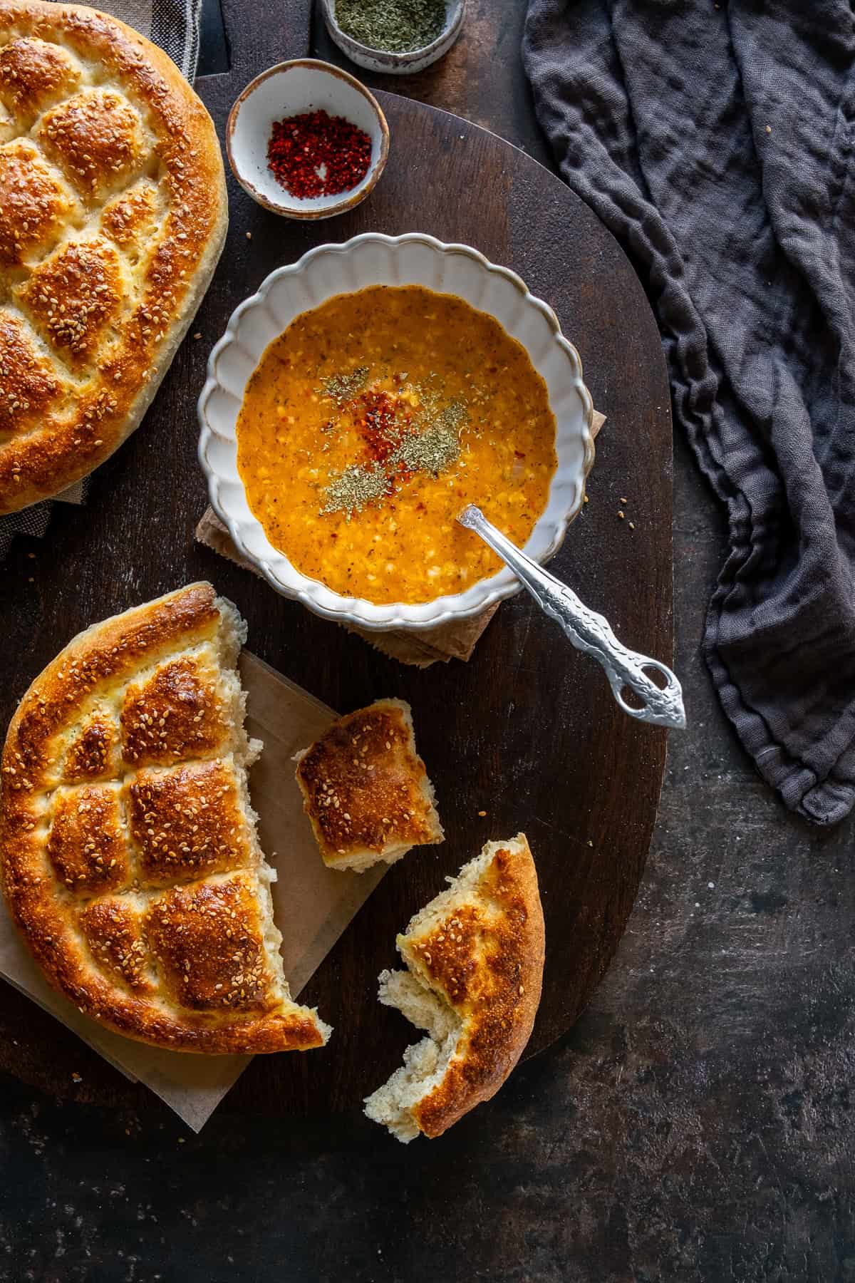 Freshly baked Turkish Pide Bread served with a bowl of Turkish soup, garnished with spices. Torn pieces of bread are placed on a wooden board, ready for dipping.