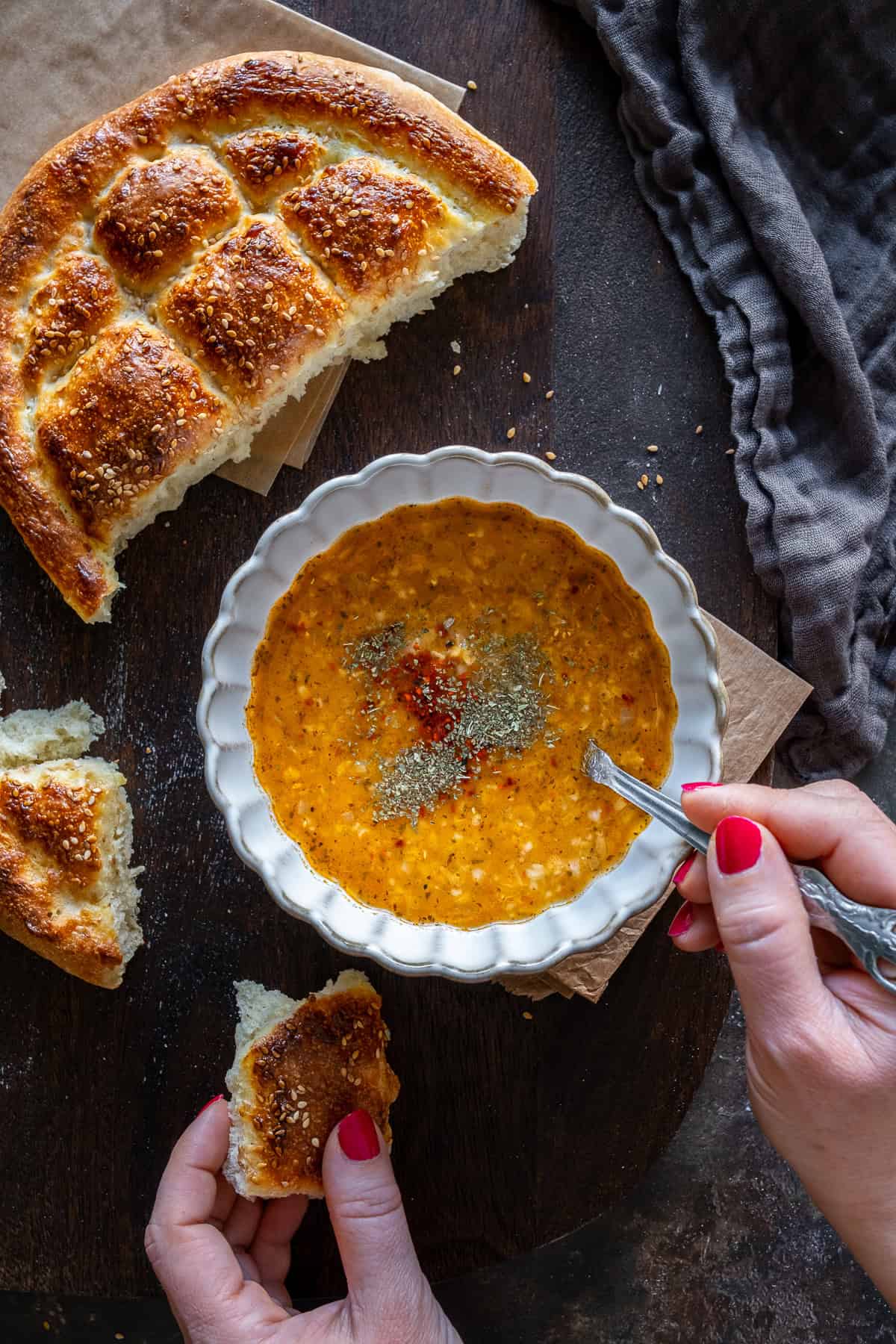 A bowl of Turkish soup garnished with spices, served with freshly baked Turkish Pide Bread. A person is holding a torn piece of bread, ready to dip.