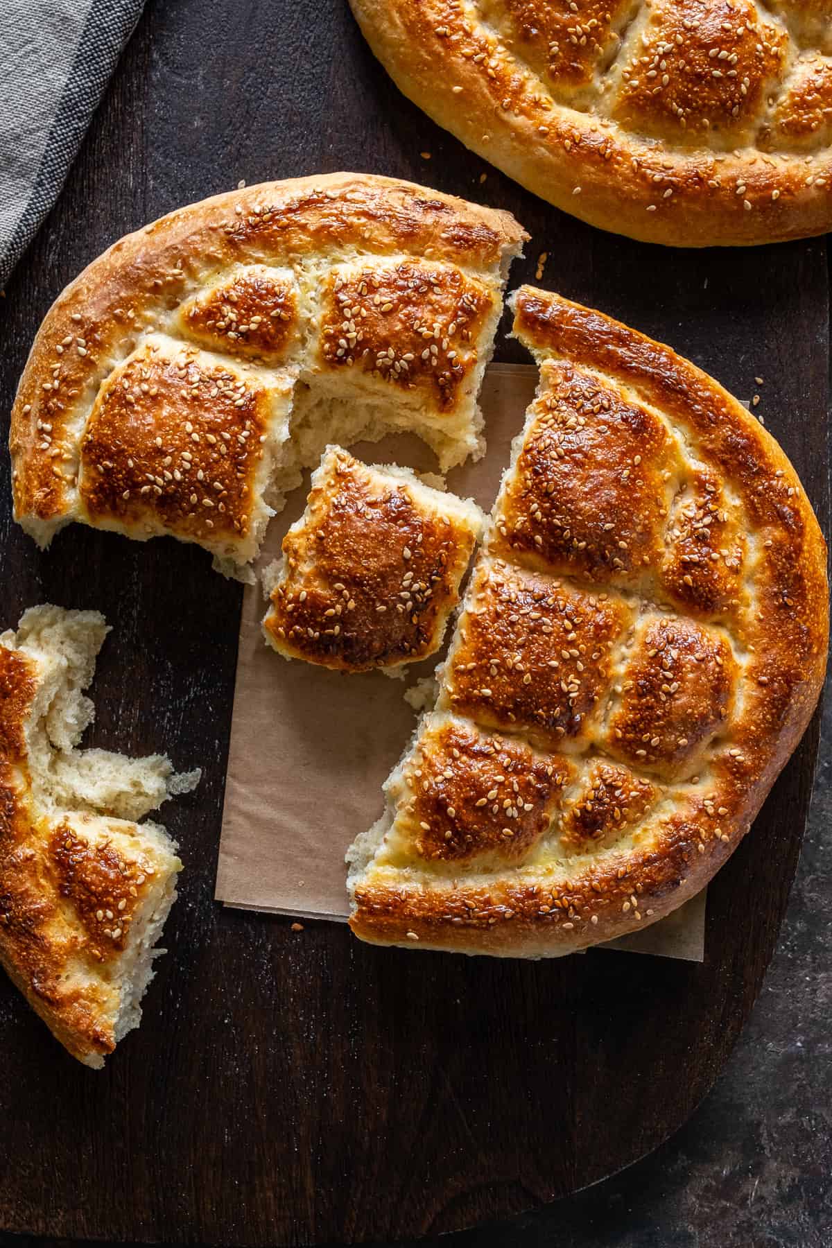 A freshly baked Turkish Pide Bread, torn into pieces, revealing its soft and fluffy interior. Another whole loaf is visible in the background.
