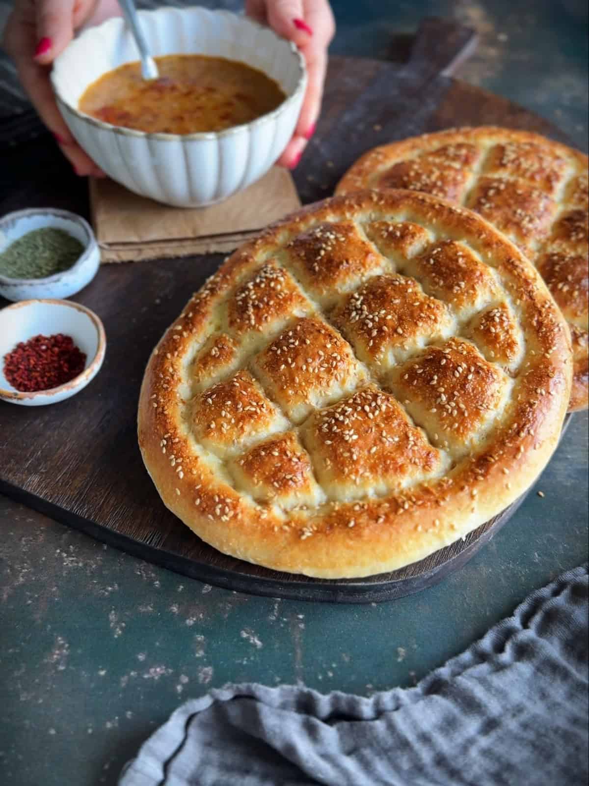 Freshly baked Turkish Pide Bread with a golden crust and sesame topping, served with a bowl of Turkish soup. Hands are seen holding the soup bowl in the background.
